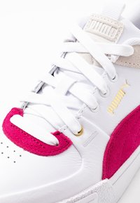 Sneaker Puma blanche avec des accents en daim rose vif, lacets blancs, languette beige et logo Puma doré sur le côté.