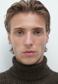 Młoda osoba z jasnobrązowymi włosami, nosząca ciemno-brązowy, dziany golf. Cechy obejmują wyraźne niebieskie oczy i gładką skórę.