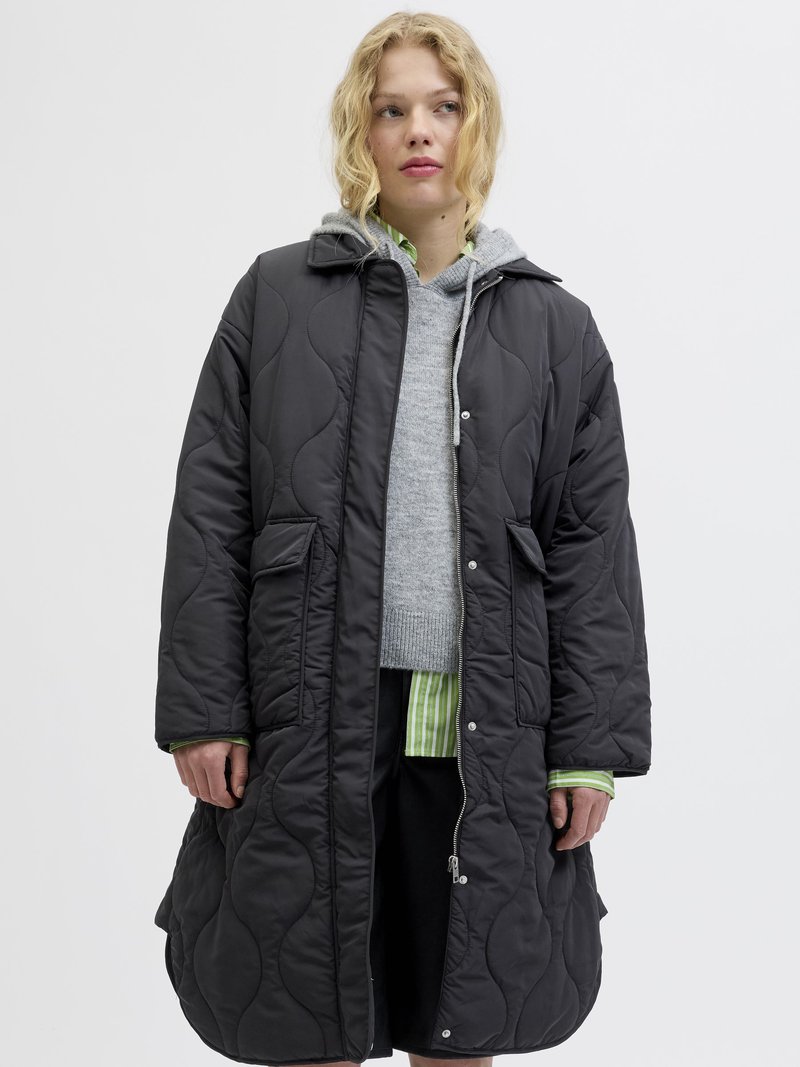 Cappotto imbottito nero con cappuccio, dotato di chiusura con zip e bottoni a pressione, e tasche laterali. Indossato sopra un maglione grigio e una camicia a righe.