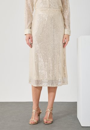 Donna vestita con una gonna midi beige in paillettes, maniche lunghe e sandali alti con cinturino dorato, che si trova su un pavimento grigio.