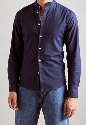 Marineblaue Langarm-Hemd mit Knopfleiste und Stehkragen, aus glattem Baumwollmaterial gefertigt, mit weißen Knöpfen und geradem Saum. Kombiniert mit blauen Jeans.
