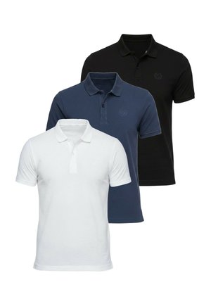 Jeune homme portant un polo bleu marine, un jean clair ample, des baskets blanches et une montre-bracelet, debout avec une main dans la poche.