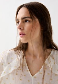 Blouse blanche avec des accents carrés dorés, tissu léger et texturé, décolleté en V et détails drapés doux, encadrée par des cheveux flottants.