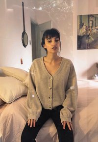Cardigan lavorato a maglia di un grigio chiaro, di grande dimensione, con grandi bottoni e polsini a coste, abbinato a pantaloni neri aderenti, contro uno sfondo di una stanza dai colori tenui.
