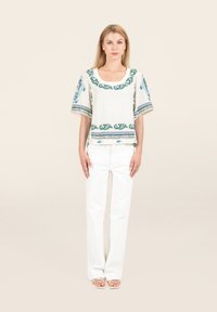 Blusa bianca ricamata con maniche corte e larghe, caratterizzata da motivi floreali blu e verdi lungo il collo e l'orlo, abbinata a jeans bianchi.