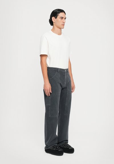Grijze spijkerbroek met patchworkstructuur, gecombineerd met een wit T-shirt zonder opschrift en zwart geborduurde instapschoenen. Model staat in profiel.