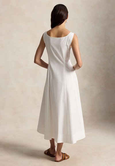 Woman with long dark hair wearing a sleeveless white midi dress and brown flat sandals, standing with hands on hips, facing away.