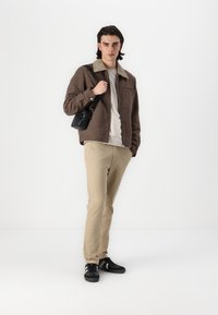 Chaqueta a cuadros marrón con cuello beige, suéter claro, pantalones beige y zapatillas negras con rayas blancas. Mochila pequeña negra al hombro.