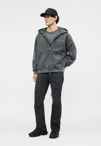 Ung mann med svart caps, iført en grå hettejakke med glidelås, slitte svarte jeans og svarte joggesko, stående foran en enkel lys bakgrunn.