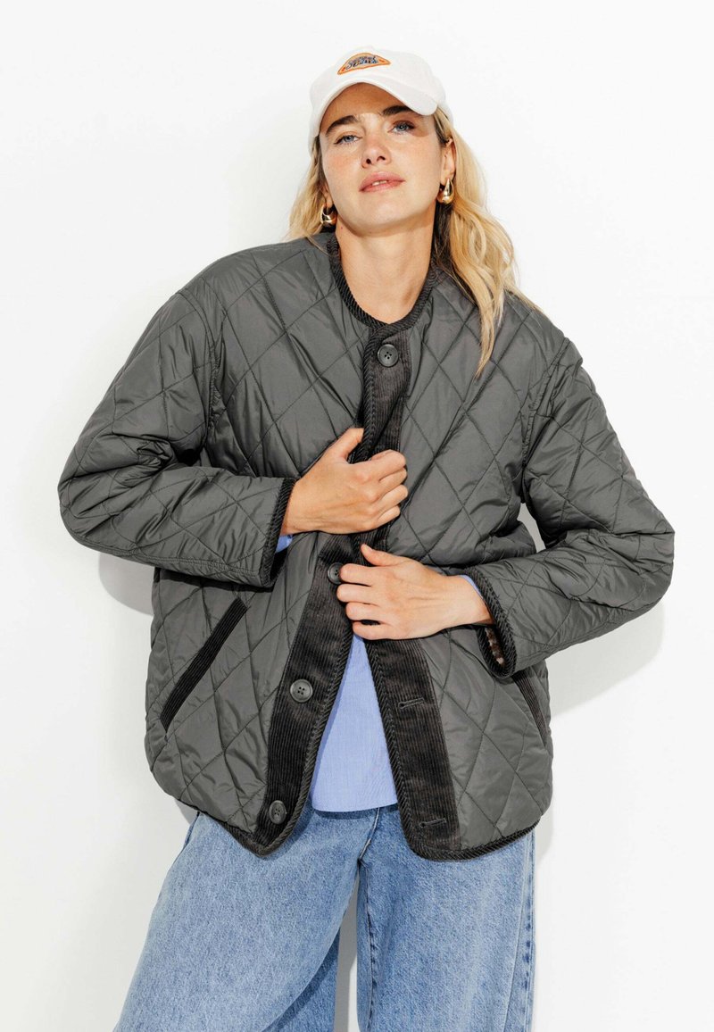 Veste grise matelassée avec un col rond, des accents en velours côtelé noir et de gros boutons. Portée sur un haut bleu clair et associée à un jean à jambes larges.