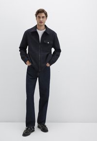 Dunkelblaues, strukturiertes Zip-Jackett mit einer Vorderentasche und weißen Knopfakzenten; kombiniert mit dunklen Jeanshosen und schwarzen Schnürschuhen.