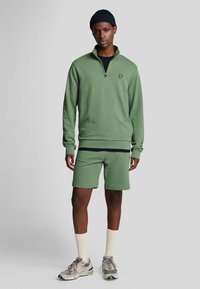 Sudadera verde con cremallera de un cuarto y logo bordado, combinada con pantalones cortos a juego. Usada con zapatillas grises y calcetines beige.