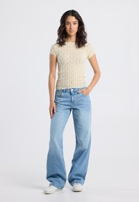 Woman with dark wavy hair wearing a short-sleeve cream lace top, light blue wide-leg jeans, white sneakers, standing with one hand in pocket.