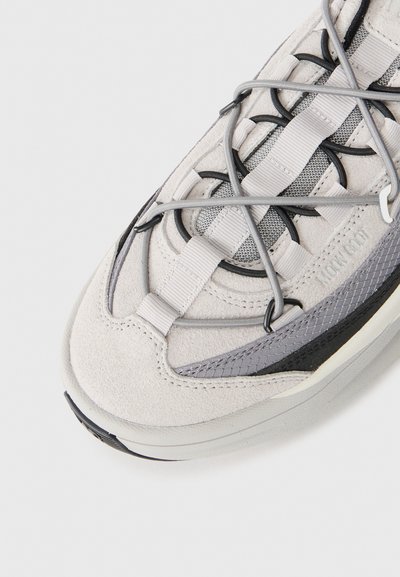 Zapatilla gris claro con materiales de ante y malla. Presenta un cordón intrincado, acentos texturizados y una suela de goma.