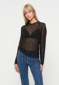Zwarte polkadot mesh top met lange mouwen en een v-hals, gemaakt van doorzichtig materiaal, gecombineerd met blauwe gestreepte high-waisted jeans.