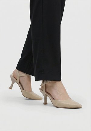 Zapatos de tacón bajo tipo kitten heel texturizados en beige con tira trasera, llevados con pantalones cortos negros sobre un fondo claro.
