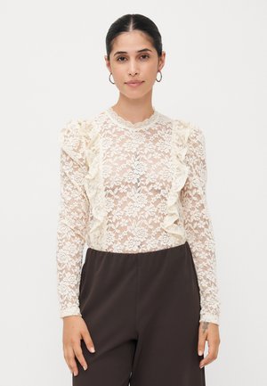 Lace blouse in ivory with ruffled accents, long sleeves, and a scalloped neckline. Worn with dark brown pants. Floral pattern throughout.