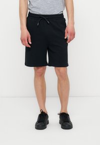 Homme portant un short noir avec cordon, un t-shirt gris rayé et des baskets noires, debout devant un fond blanc.
