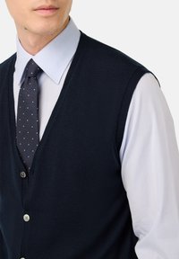 Jūras spēku adīta veste ar V veida kaklu, apģērbu no tekstūrēta auduma, uzvilkta virs gaiši zila krekla un tumšas punktainas kaklasaites. Redzams piecu pogu aizdare.