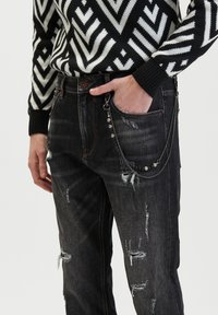 Zwarte denim jeans met gescheurde details en een zilveren kettingaccessoire dat aan de zak is bevestigd, gecombineerd met een zwart-wit gestreepte trui.