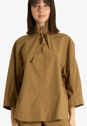 Donna con capelli scuri e lisci che indossa una maglia marrone larga con maniche a tre quarti e apertura del colletto annodata.