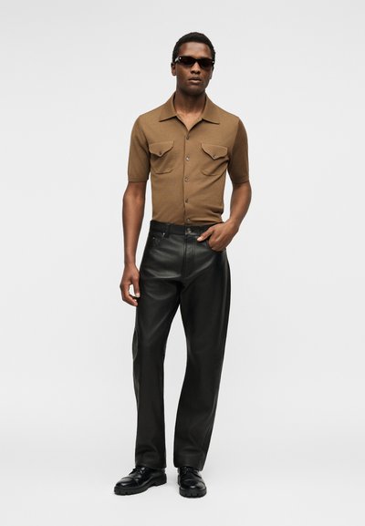 Άνδρας που φορά καφέ κοντομάνικο πουκάμισο με κουμπιά, μαύρο δερμάτινο παντελόνι, μαύρα παπούτσια και σκούρα γυαλιά ηλίου, στέκεται μπροστά σε απλό φωτεινό φόντο.