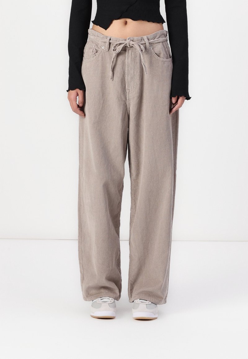 Personne portant un pantalon large en velours côtelé beige avec une taille à cordon, un top noir court à manches longues et des baskets blanches.