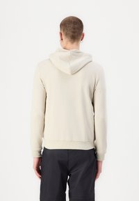 Beige kapucnis pulóver, húzózsinórral, bordázott mandzsettákkal és aljjal. Puha tapintású, laza fazonú és minimalista dizájnú. Sötét sorttal viselve.