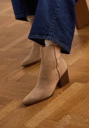 Beige Ankle-Boots aus Wildleder mit Blockabsätzen, getragen zu blauen Jeans und beigen Socken auf einem Holzparkettboden.