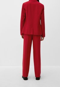 Costume pantalon rouge avec une veste cintrée et un pantalon large. Tissu lisse, lignes épurées, sans motifs ni embellissements visibles.