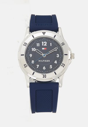 Marine rubber band horloge met een zilveren roestvrijstalen kast, donkerblauwe wijzerplaat, grote witte cijfers en blauwe accenten op de wijzers. Model: Hilfiger.