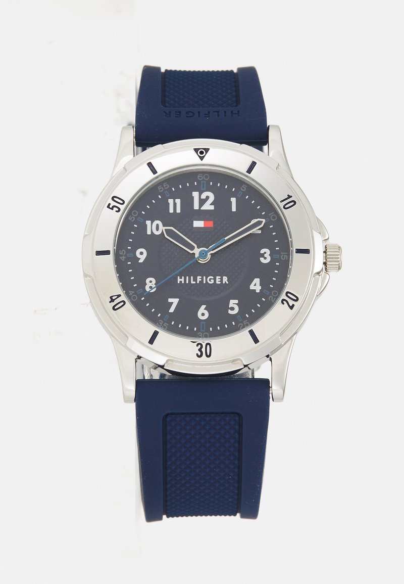 Relógio com pulseira de borracha azul-marinho, caixa de aço inoxidável prateado, mostrador azul escuro, grandes números brancos e ponteiros com detalhes azuis. Modelo: Hilfiger.