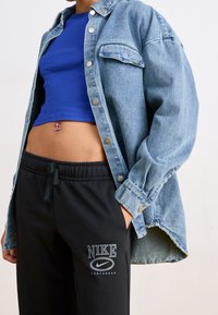 Lichtblauwe denim shirt met een ontspannen pasvorm, zilveren knoopdetails, gedragen over een aangesloten koninklijkblauw crop top en zwarte Nike joggingbroek.