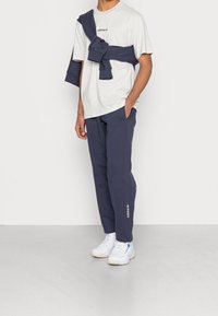 Vit bomullströja med en mörk marinblå sweatshirt knuten runt axlarna, tillsammans med mörk marinblå sweatpants och vita sneakers.