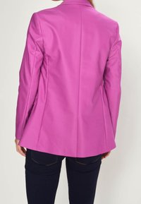 Růžový blejzr s dobře padnoucím střihem a hladkou texturou, s klopovým límcem a zadním rozparkem. Kombinovaný s tmavými džíny.