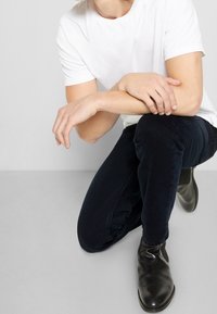 Vit bomull t-shirt, mörkblå denimjeans och svarta läderankelboots. Modell på knä med armarna vilande på det böjda knät. Enkelt design.