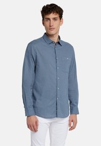Camicia azzurro chiaro a maniche lunghe in un tessuto testurizzato, con chiusura a bottoni e un'unica tasca sul petto. Indossata con pantaloni bianchi.