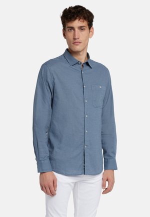 Chemise à manches longues bleu clair en tissu texturé, avec un design à boutons et une poche poitrine unique. Portée avec un pantalon blanc.