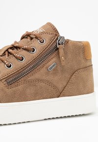 Bottine en daim marron avec une semelle en caoutchouc blanche, équipée d'une fermeture éclair latérale et de lacets assortis, marquée d'un logo argenté.