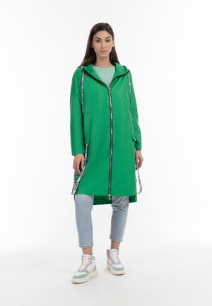 Groene lichtgewicht regenjas met een capuchon, voorzien van een ritssluiting aan de voorkant en zijzakken, gecombineerd met lichtblauwe jeans en multicolore sneakers.