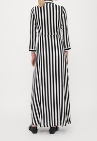 Kobieta w długiej czarno-białej sukience w pionowe paski z rękawami o długości trzy czwarte, stojąca tyłem, w czarnych płaskich butach.
