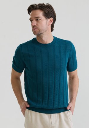 Mand med kort brunt hår iført en turkis tekstureret kortærmet sweater og lyse bukser, kigger til siden med hænderne i lommerne.