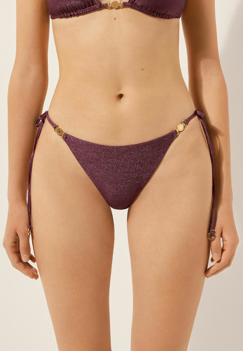 Femme portant un bas de bikini violet scintillant avec des liens sur les côtés et des accents circulaires dorés, montrant le ventre et le haut des cuisses.
