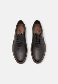 Zapatos de cuero negro con un acabado suave, que presentan un panel de tela texturizada, cordones redondos y una plantilla marrón claro con el logotipo de la marca.