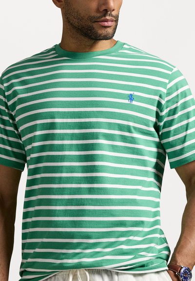 Tricou pe fundal verde și alb, cu dungi, fabricat din bumbac, cu guler rotund. Are un mic logo albastru pe piept și mâneci scurte.