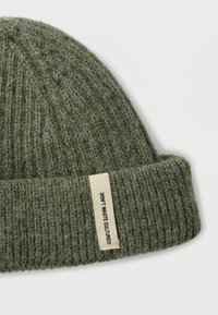 Πράσινος πλεκτός σκούφος με ραβδώσεις και χοντρό μανσέτα, με ορθογώνια ετικέτα που γράφει "ΜΗ ΣΠΑΤΑΛΑΣ ΚΟΥΛΤΟΥΡΑ." Ανθεκτικό ύφασμα με υφή.