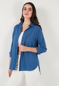 Camisa azul de botones hecha de un tejido texturizado, con cuello, mangas largas y dobladillo curvo. Usada sobre una blusa blanca y combinada con pantalones blancos.