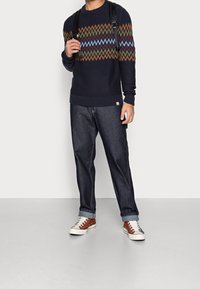 Pull marin avec motif multicolore en zigzag, jean en denim et baskets marron avec accents blancs. Look décontracté avec un fond neutre.