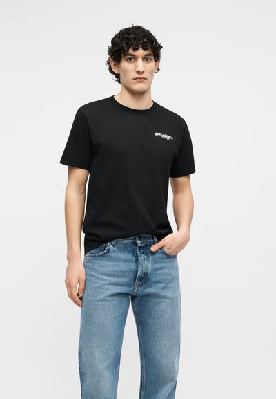 Homme aux cheveux bouclés portant un t-shirt noir Off-White et un jean bleu, debout avec une main dans la poche sur un fond uni.