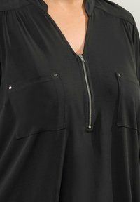 Svart blus med dragkedja i halsen, två framfickor och ett lätt material. Har en plisserad design vid axlarna.